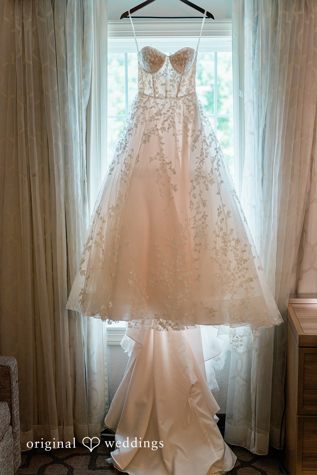 Detailed wedding dress hanging elegantly in front of a bright window.