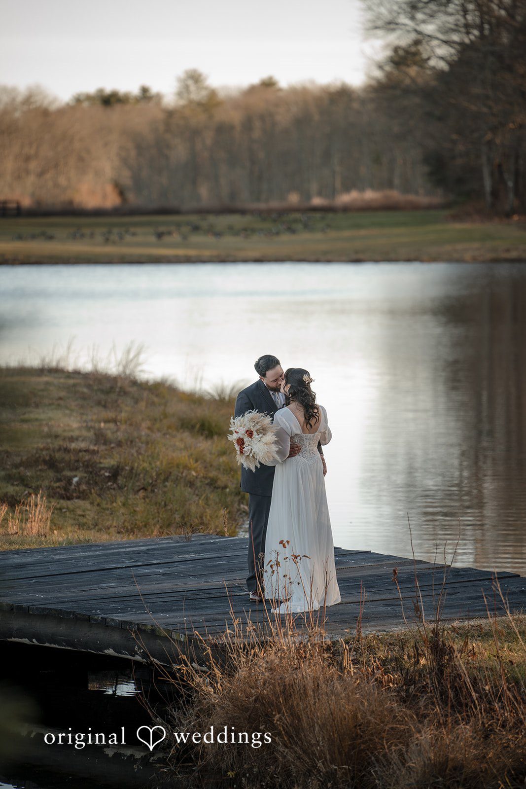 Carolyn + Robert Bride and groom embracing beside a peaceful lakeside landscape.