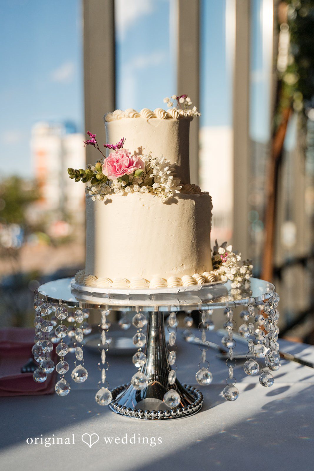 Two-tier wedding cake with floral accents displayed on crystal stand at Gather at Deer District
