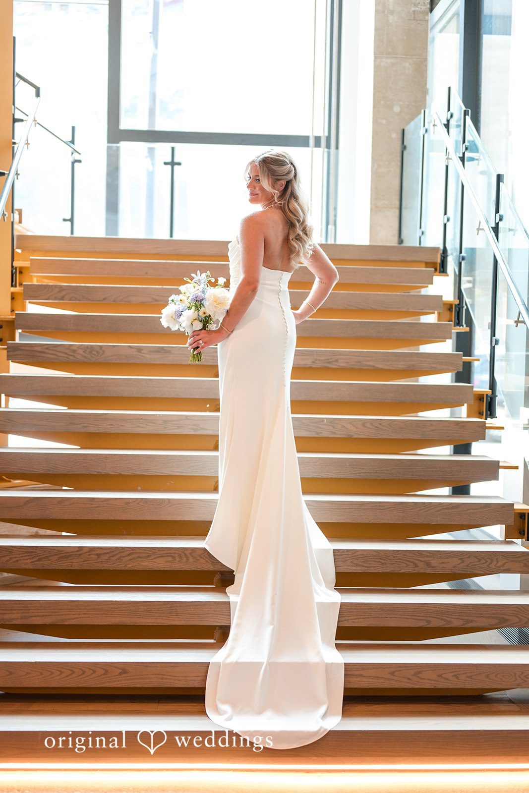 Bride in elegant fitted gown posing on staircase with bouquet before ceremony