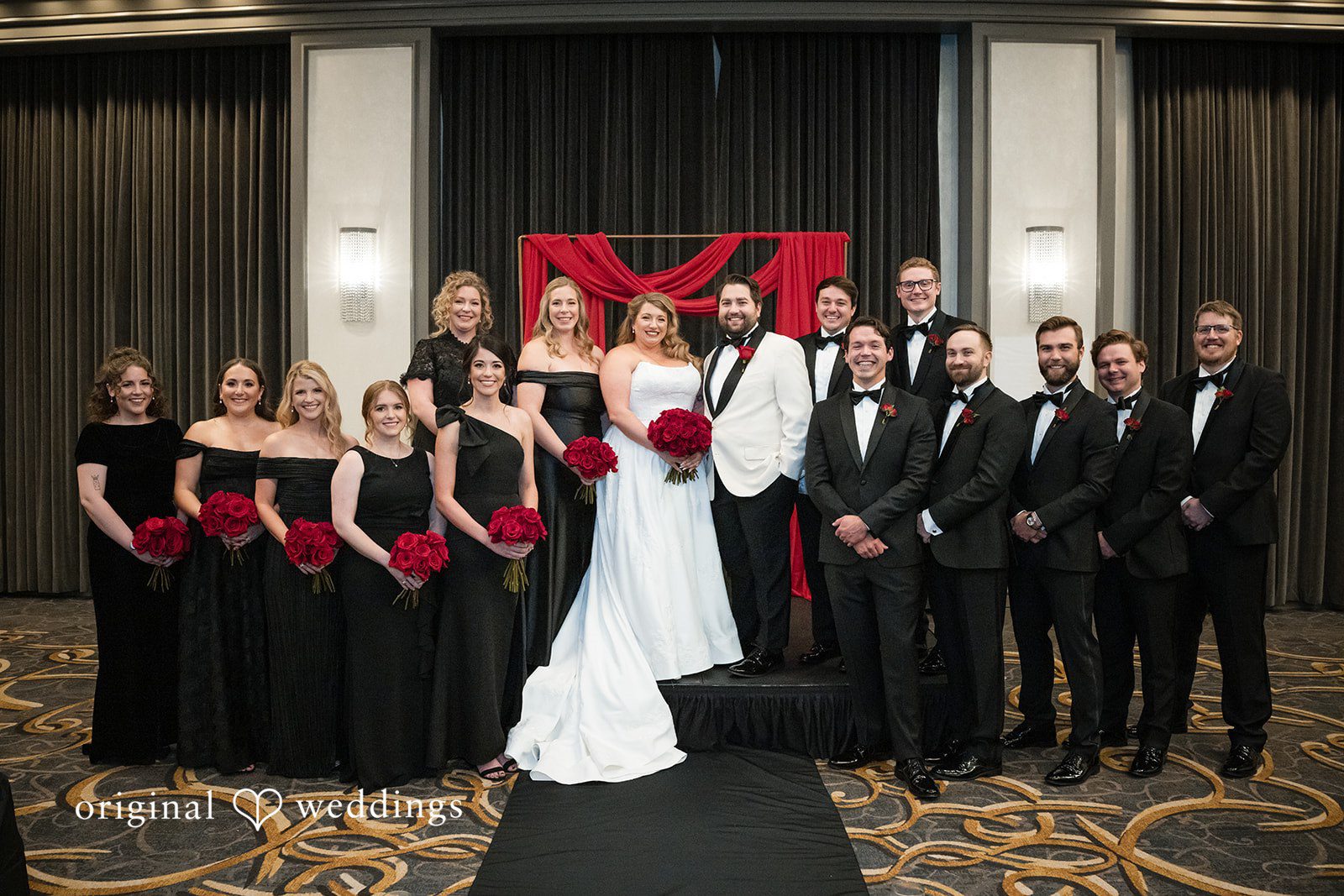 Large wedding party posing together, with bridesmaids in black dresses and groomsmen in black tuxedos.