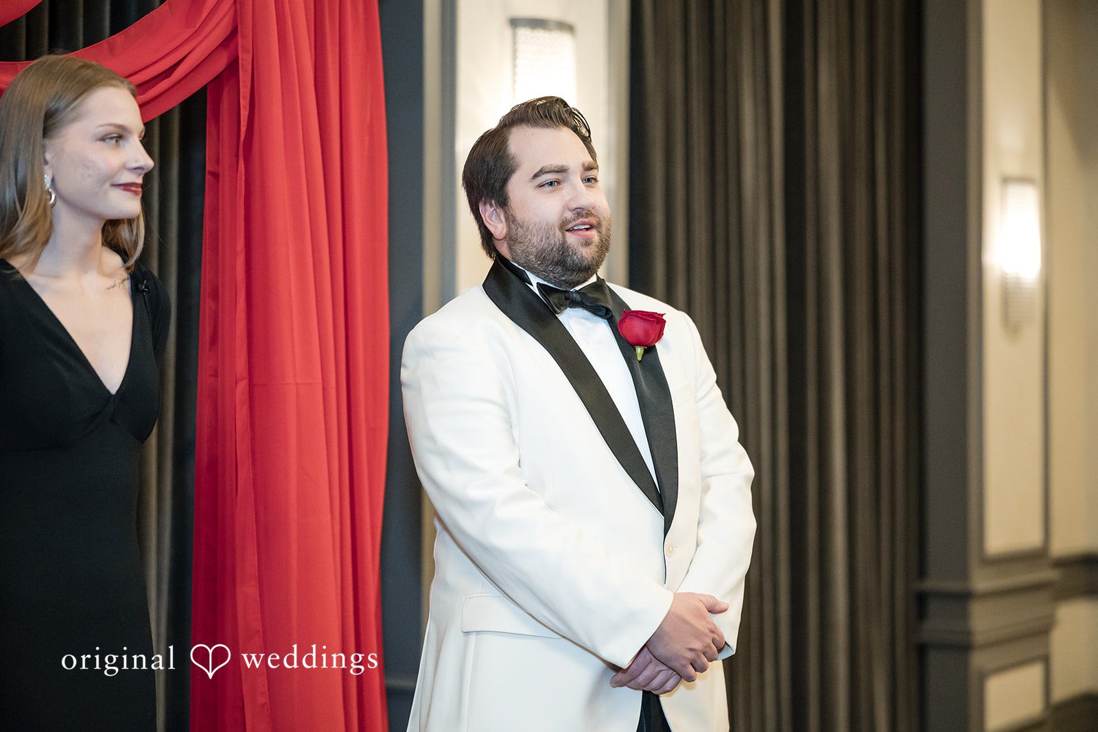 Groom in a white tuxedo jacket with black lapels standing near a red curtain.