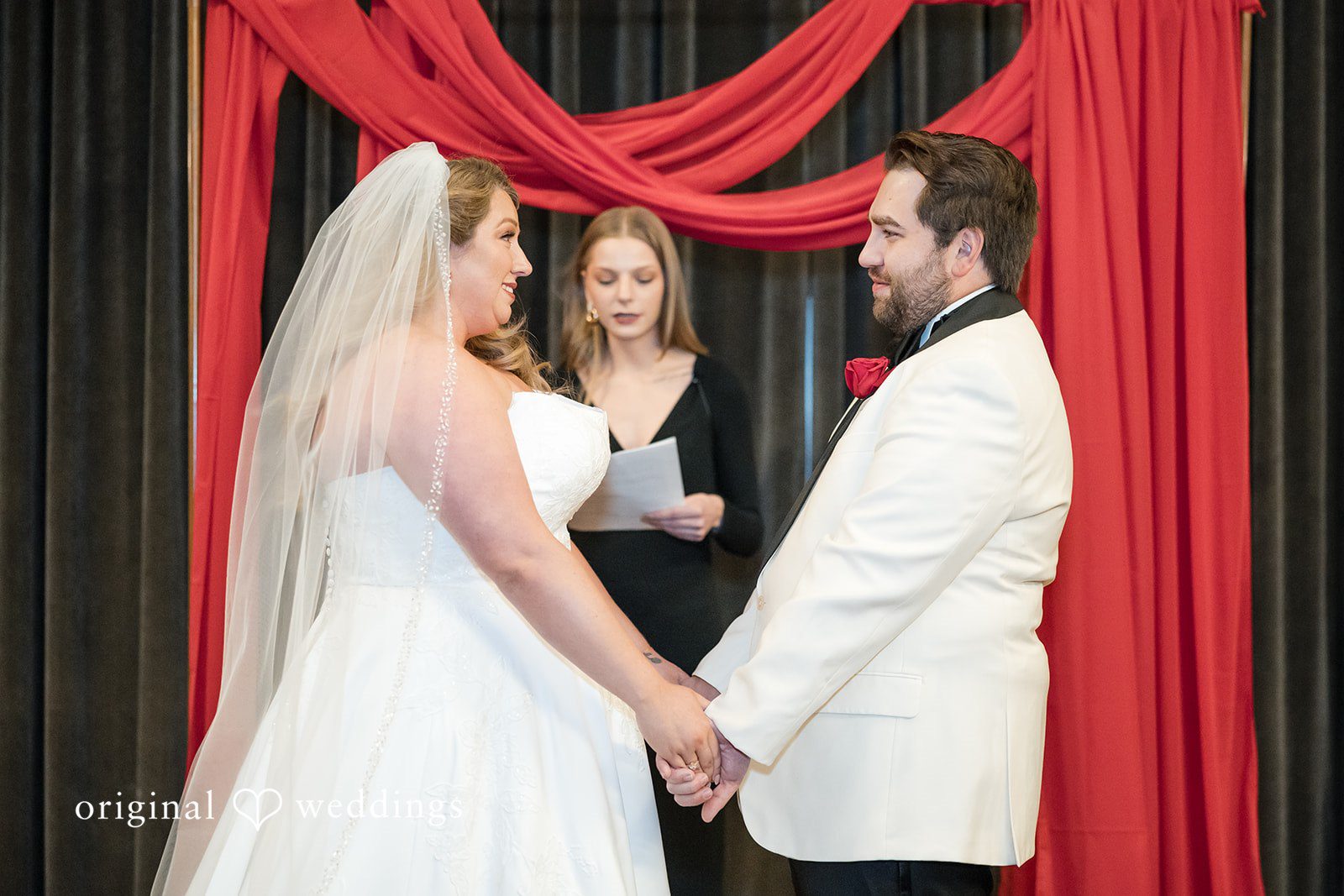 Bride and groom holding hands during the wedding ceremony in front of red drapes.