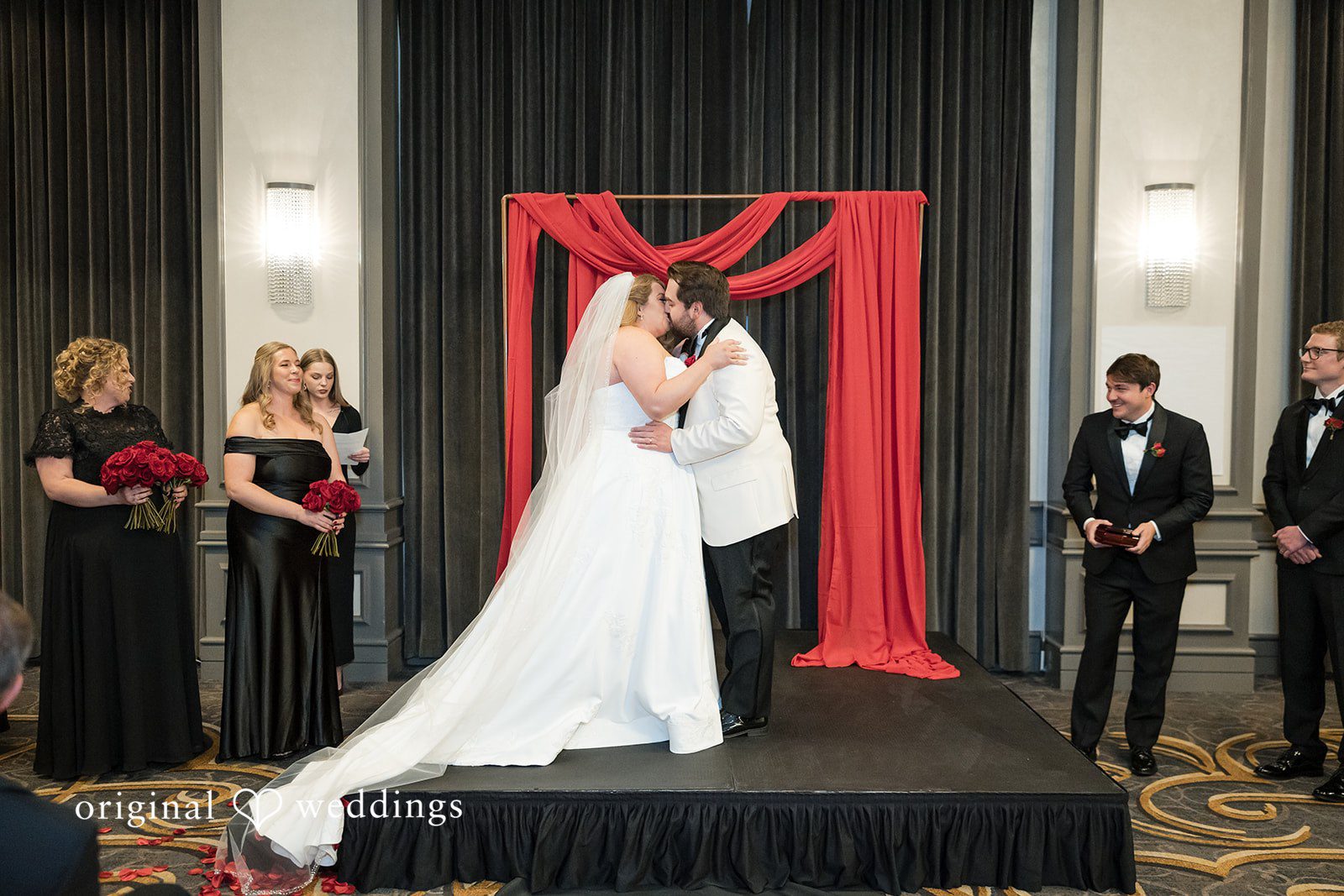 Bride and groom sharing a hug or kiss on a stage decorated with red fabric.