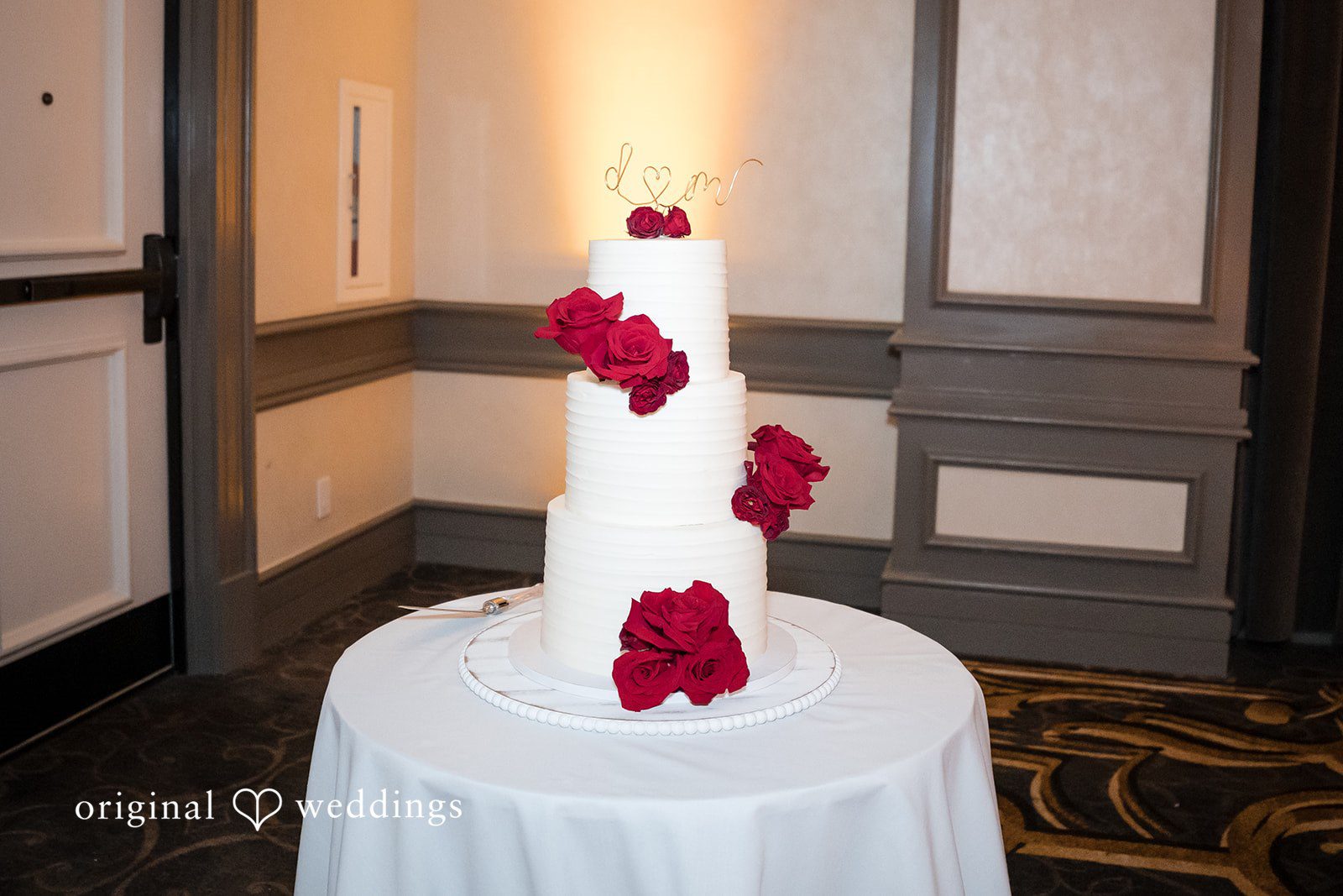 Three-tiered white wedding cake decorated with red roses on a round table.