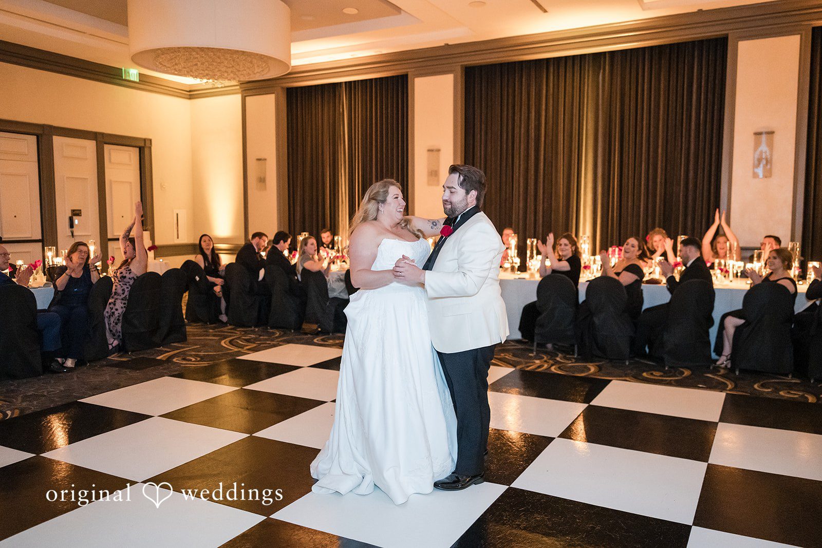 Wide shot of the bride and groom dancing on a checkered black-and-white dance floor surrounded by reception tables.