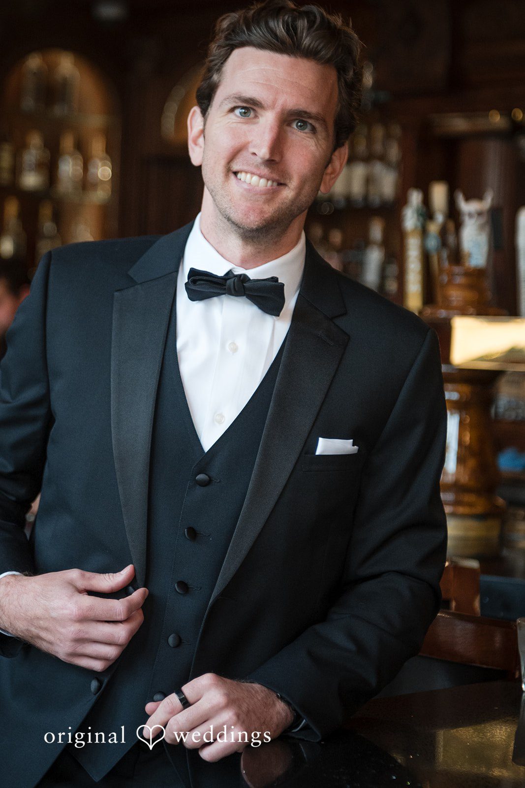 Tabatha + Justin A smiling groom poses in a classic black tuxedo and bowtie.