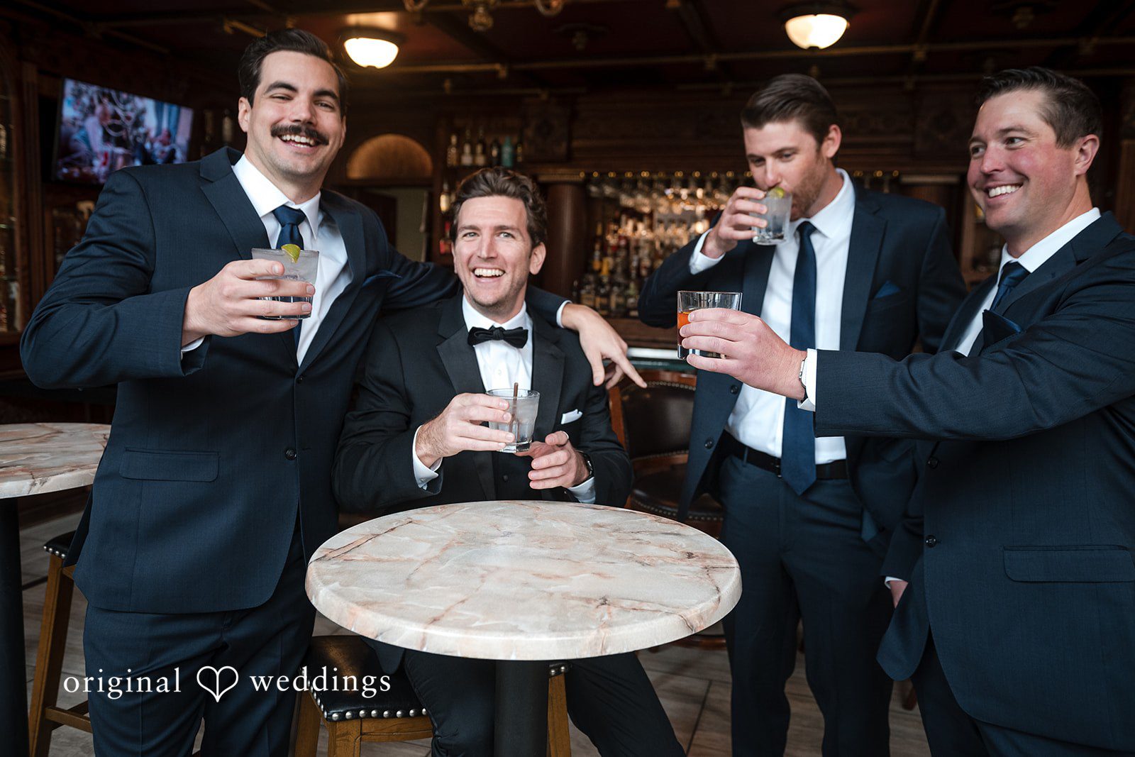 Tabatha + Justin Men in suits and tuxedos laugh and toast drinks around a round table.