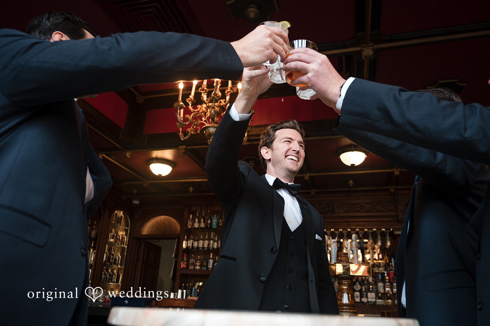 Tabatha + Justin Hands raise glasses to make a toast above a smiling groom in a tuxedo.