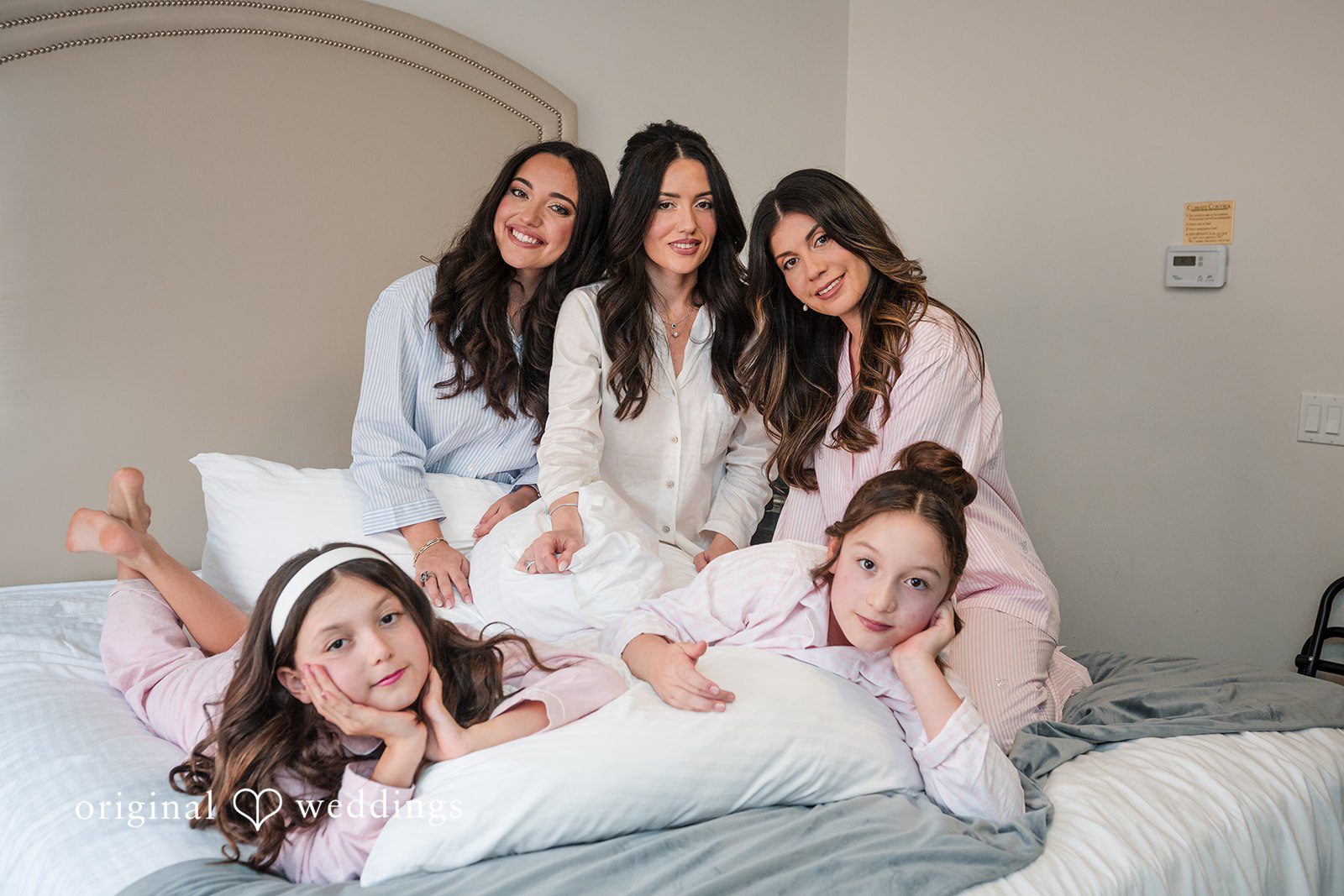 Tabatha + Justin Bride with Bridesmaid wearing light pink robes pose together playfully on a bed.