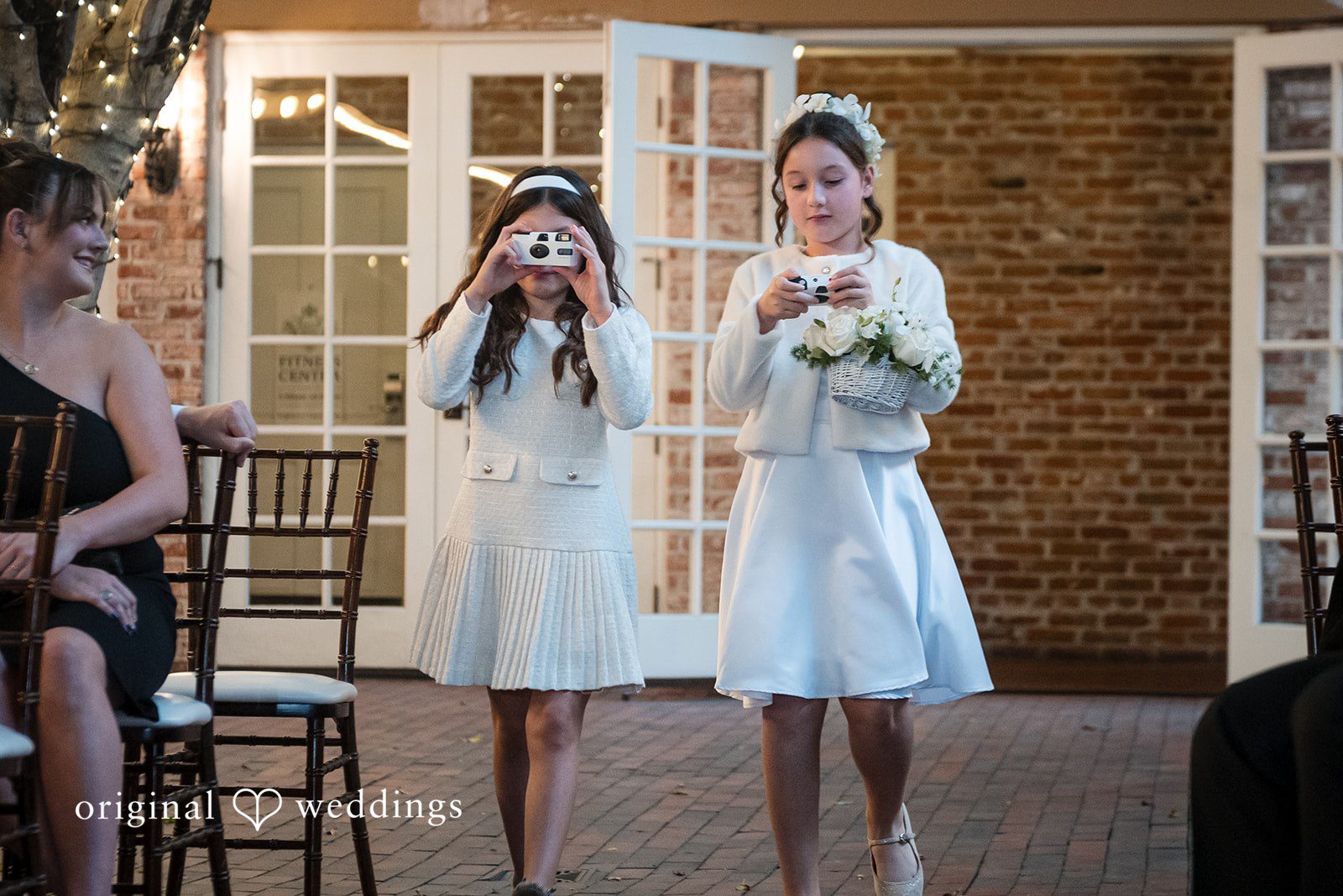 Tabatha + Justin Young flower girls in white dresses walk down the aisle holding baskets.