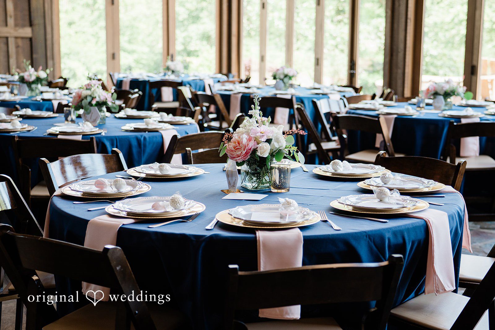 Round reception table set with a navy tablecloth, pink napkins, gold chargers, and floral centerpieces.