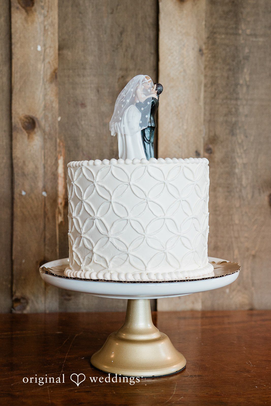 White two-tiered wedding cake with a textured pattern texture sitting on a gold stand.