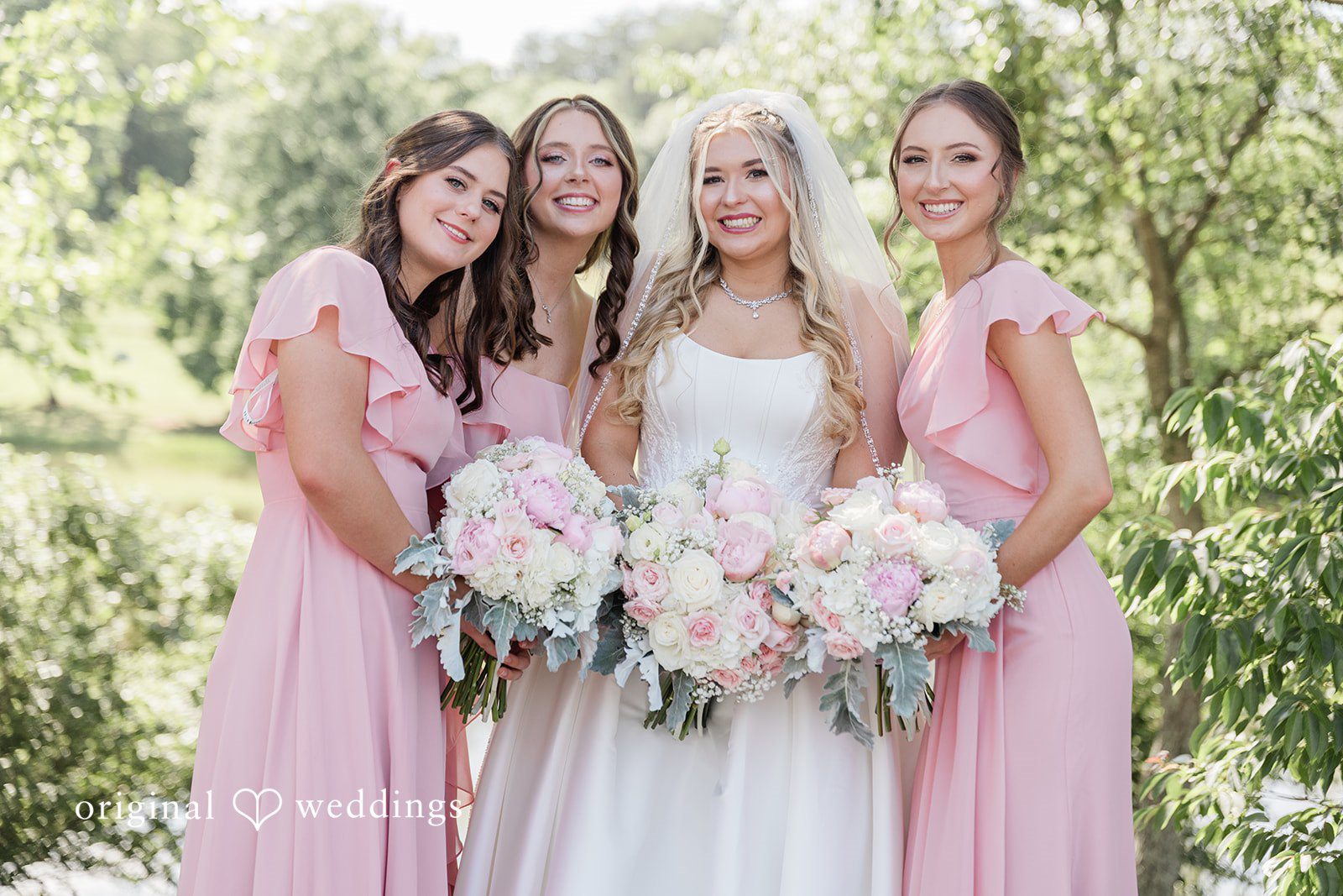 Bride holding a large pastel bouquet while posing with three bridesmaids in matching light pink dresses.