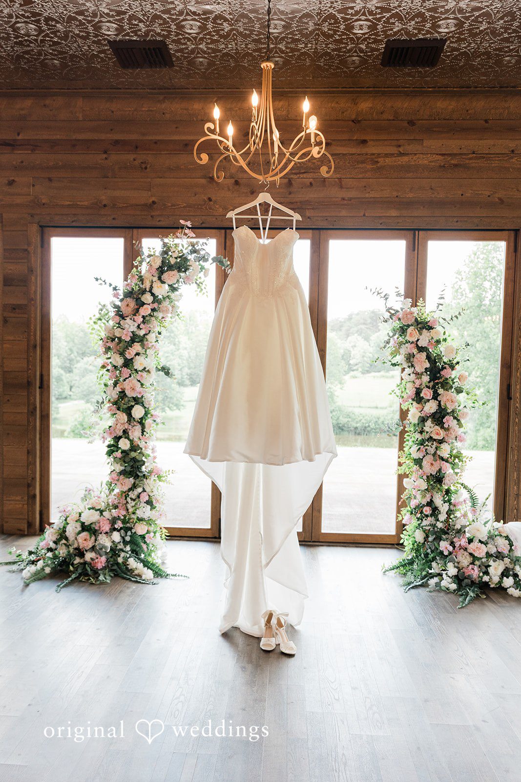 Wedding dress hanging under a chandelier in a wooden room flanked by two floral arches.