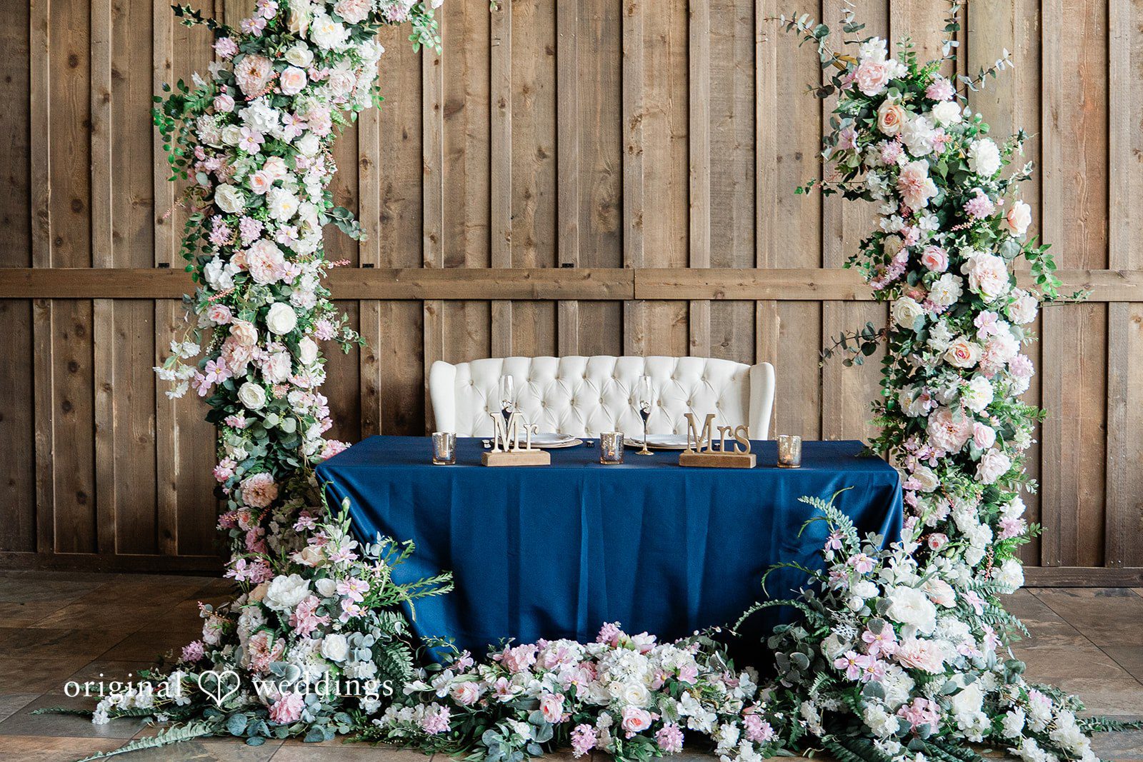 Sweetheart table draped in navy blue fabric beneath a tall floral archway with greenery.