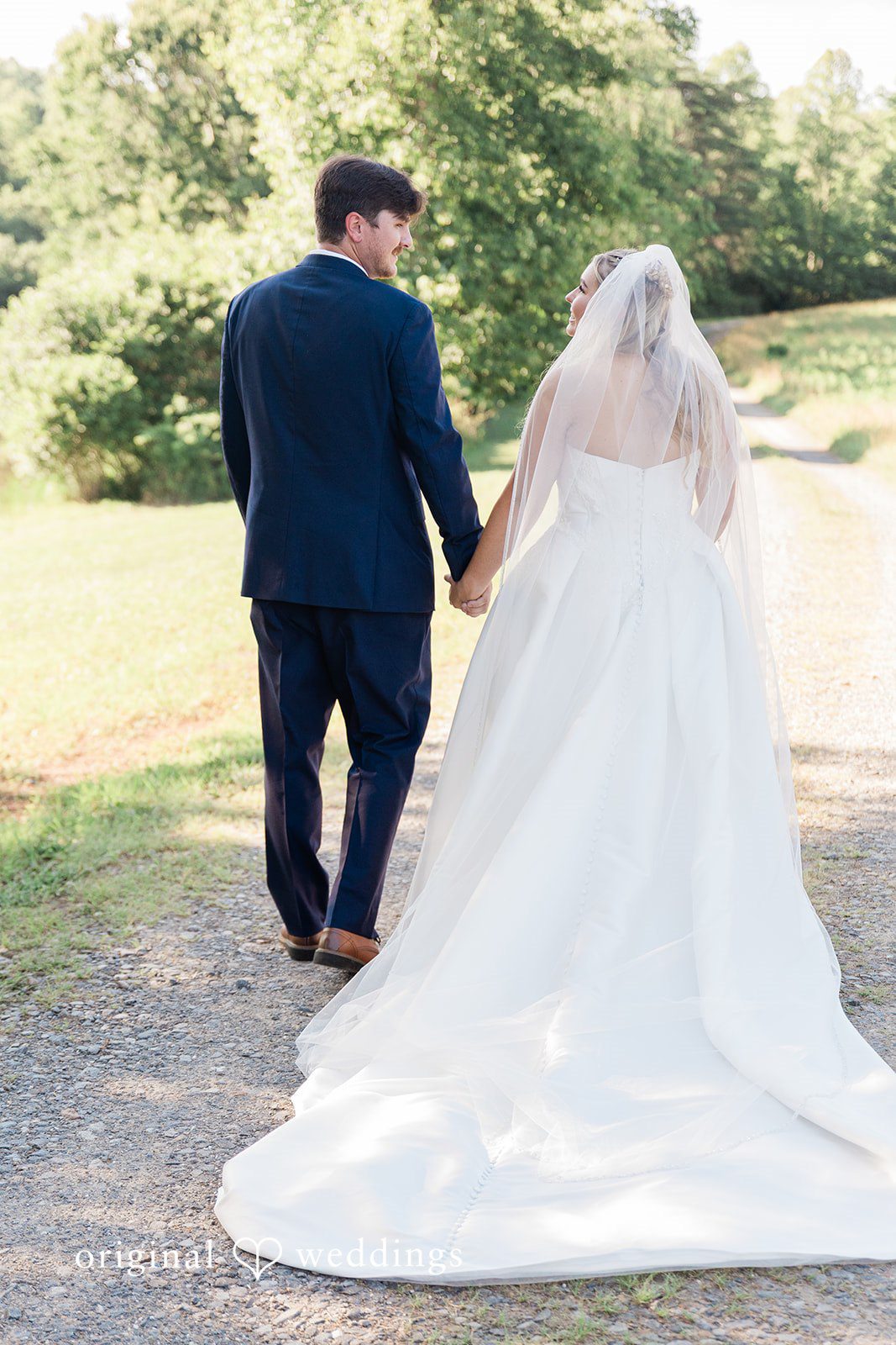 Rear view of a bride and groom holding hands while walking along a grassy path.