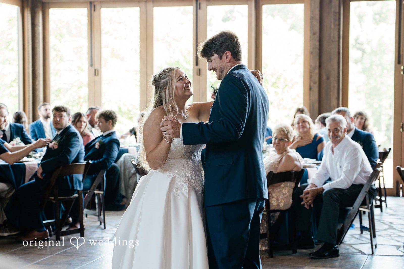 Bride and groom sharing their first dance together on a wooden floor in front of seated guests.