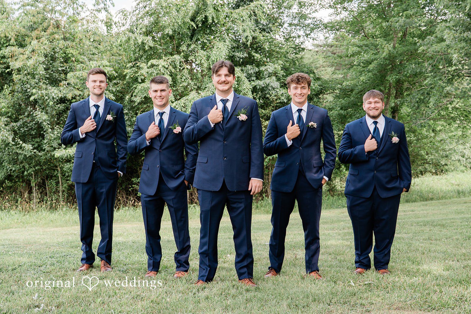 Groom and groomsmen in matching navy suits holding drinks and posing together on a lawn.
