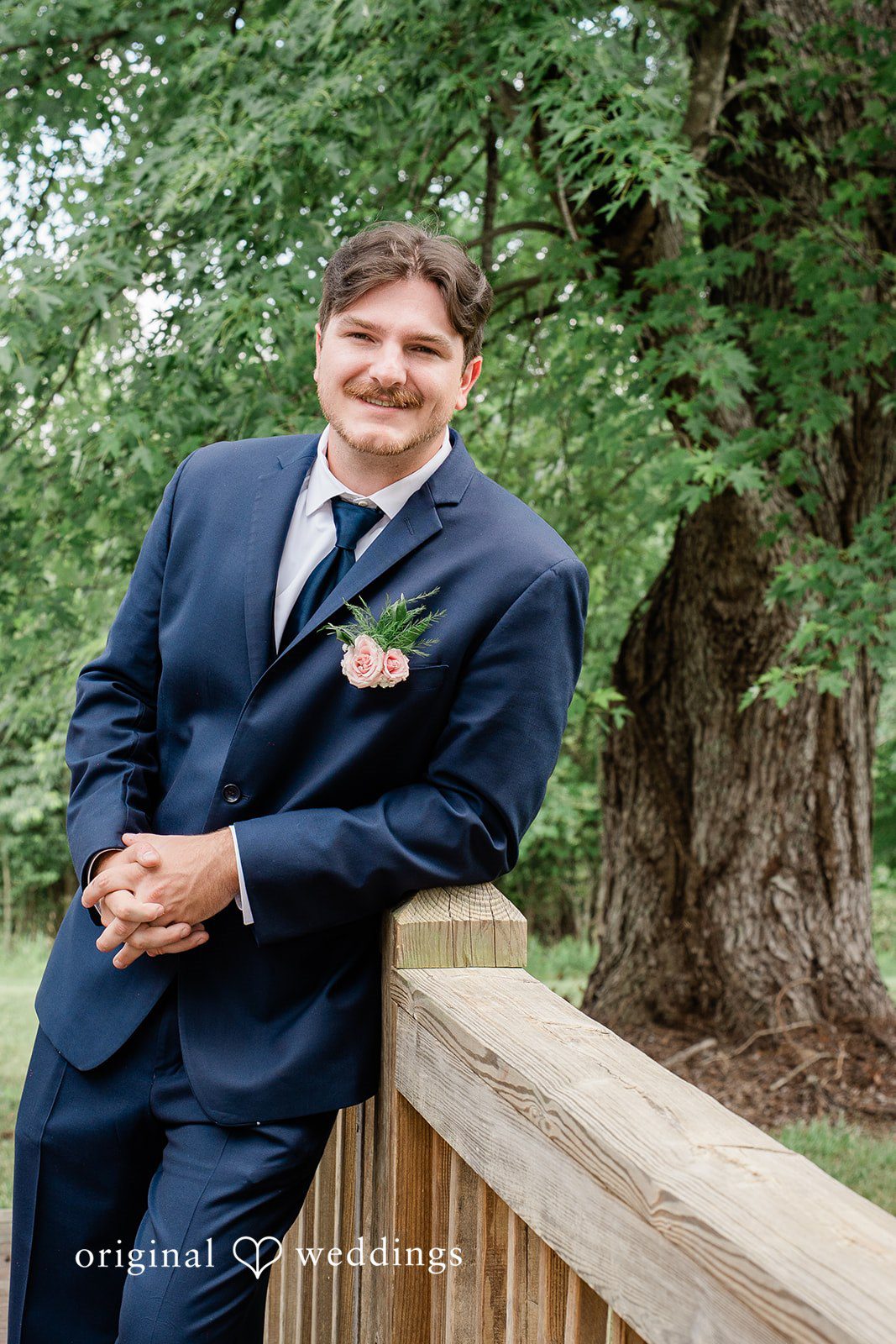 Groom in a navy blue suit smiling while leaning against a wooden bridge railing.