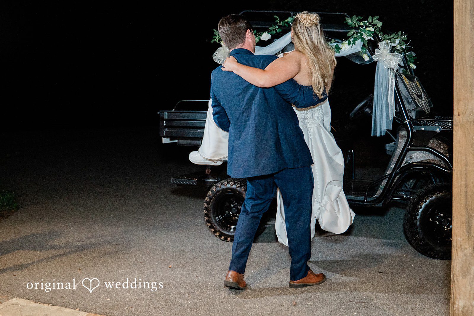 Groom carrying his bride in his arms toward a decorated golf cart for their wedding exit.