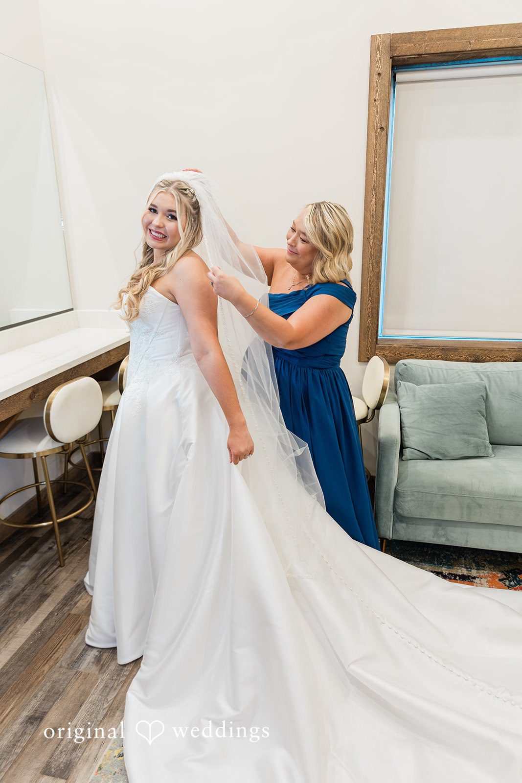 oman in a blue dress helping a bride adjust the train of her white wedding gown.
