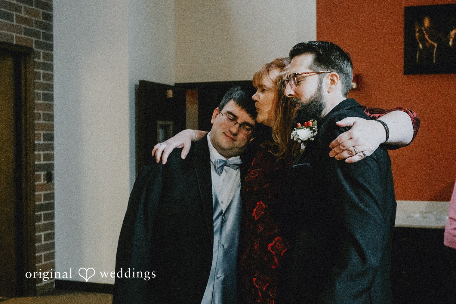 Daniel + Amanda A woman stands between two men, hugging the groom on the right and another man in a suit.