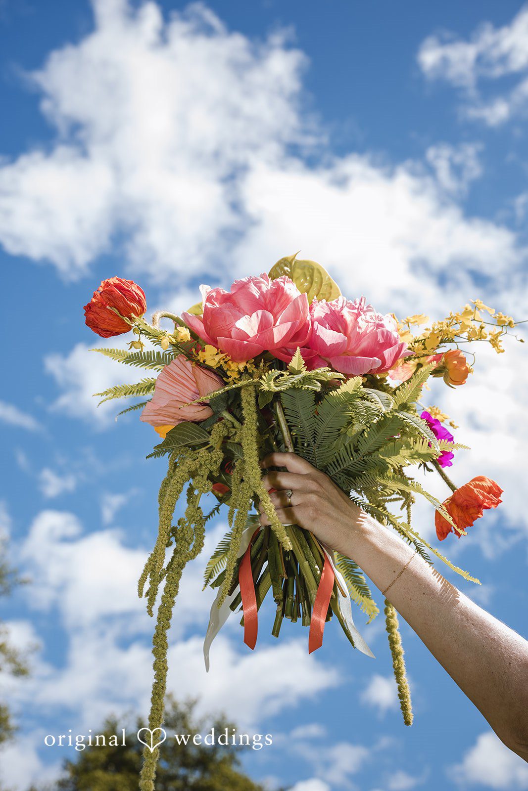 Benjamin + Amber A vibrant bridal bouquet with pink and orange blooms held against a bright blue sky at Outlook Inn