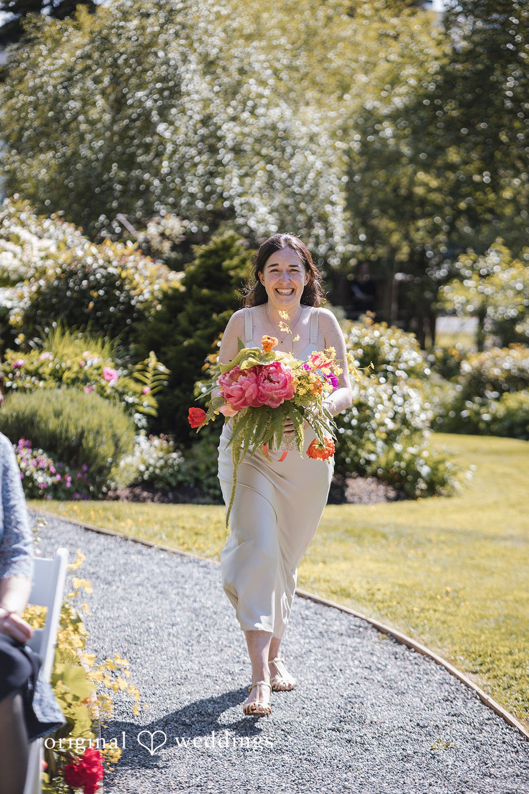 Benjamin + Amber A joyful bride walks down the garden path holding a vibrant bouquet