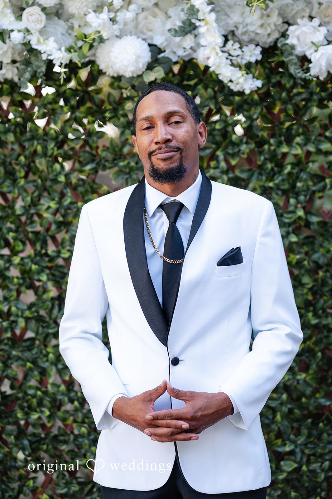 A groom wearing a white tuxedo jacket with black lapels posing in front of a green hedge.