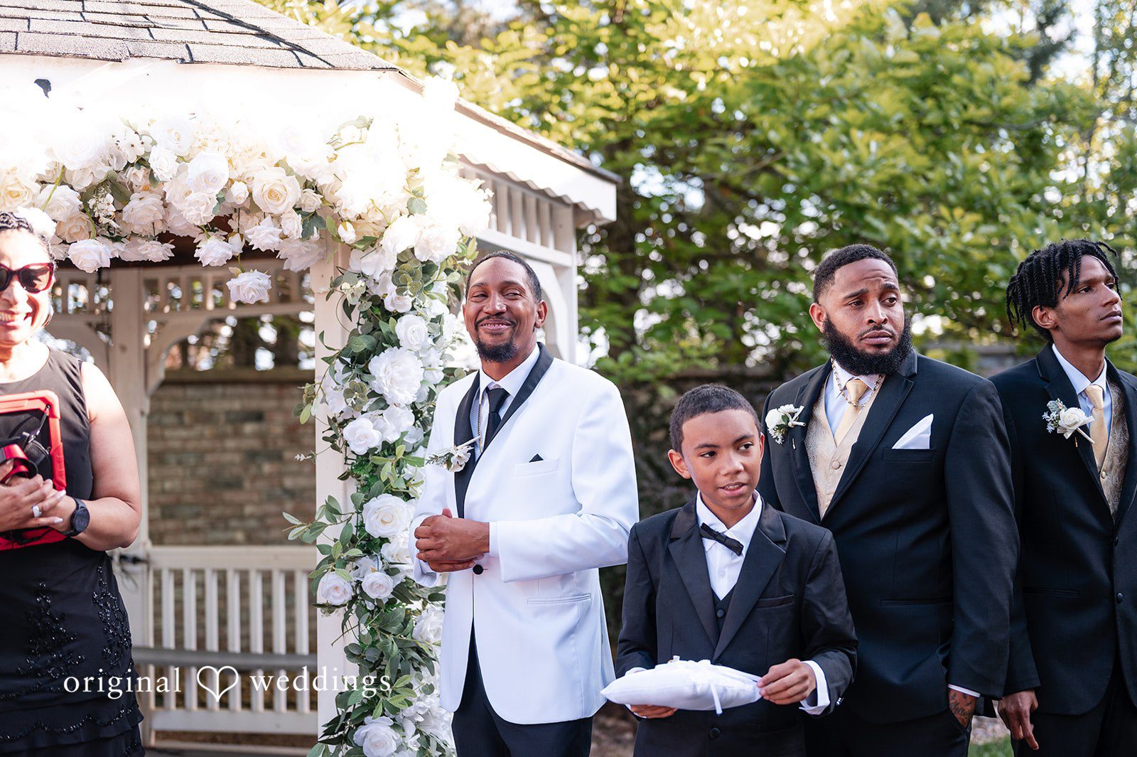 A groom in a white tuxedo and a young boy holding a ring pillow standing at a wedding ceremony.