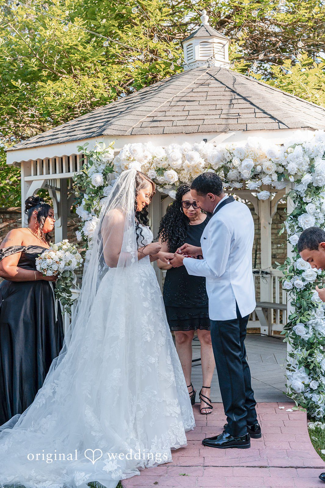 A bride and groom exchanging wedding rings in front of a floral arch and gazebo.