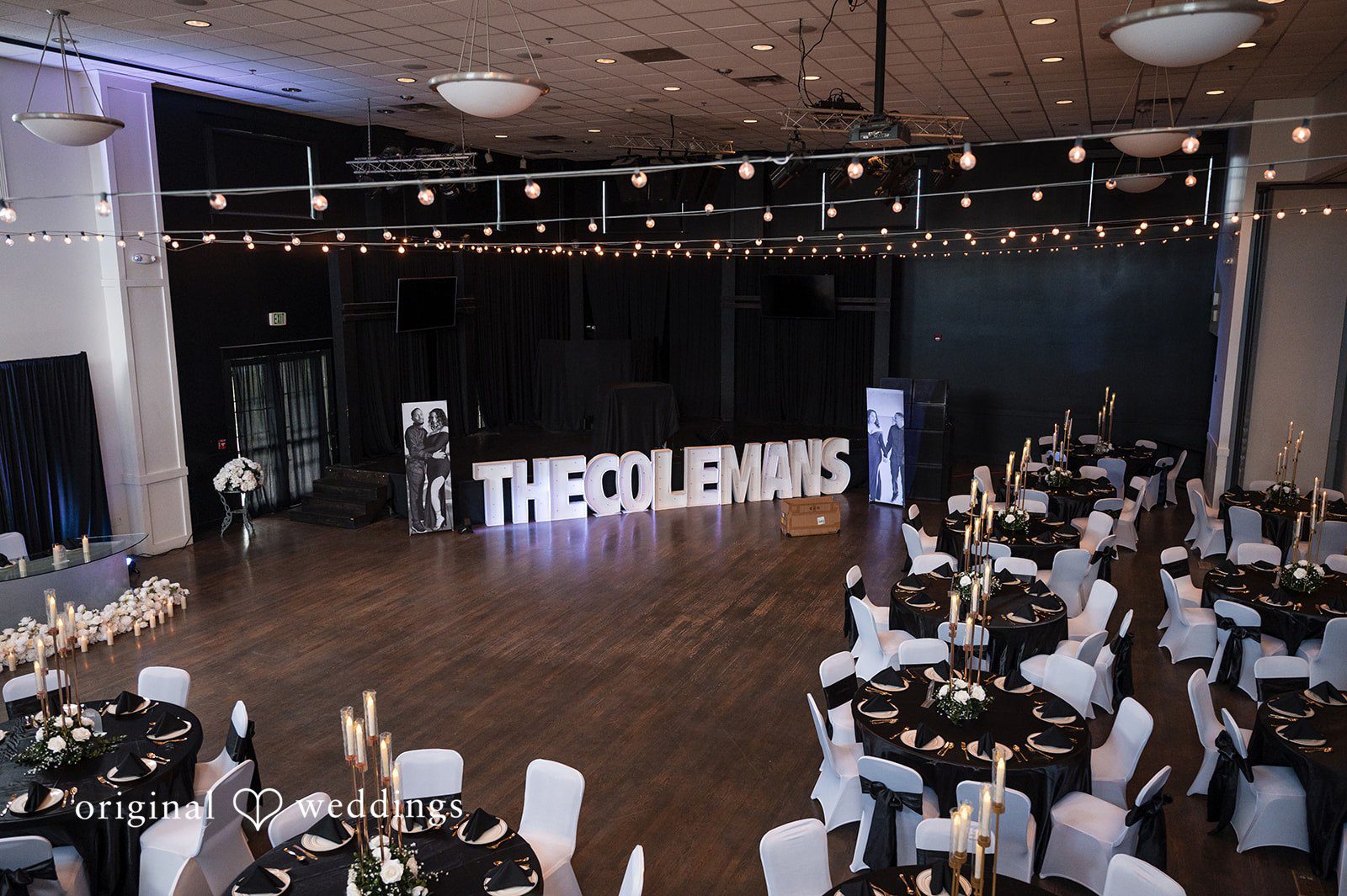 A wedding reception hall decorated with dark tables, fairy lights, and a custom lit-up sign.