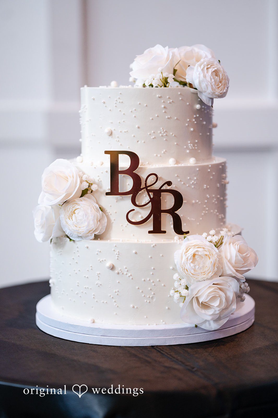 A three-tiered white wedding cake decorated with flowers and the initials "B&R".