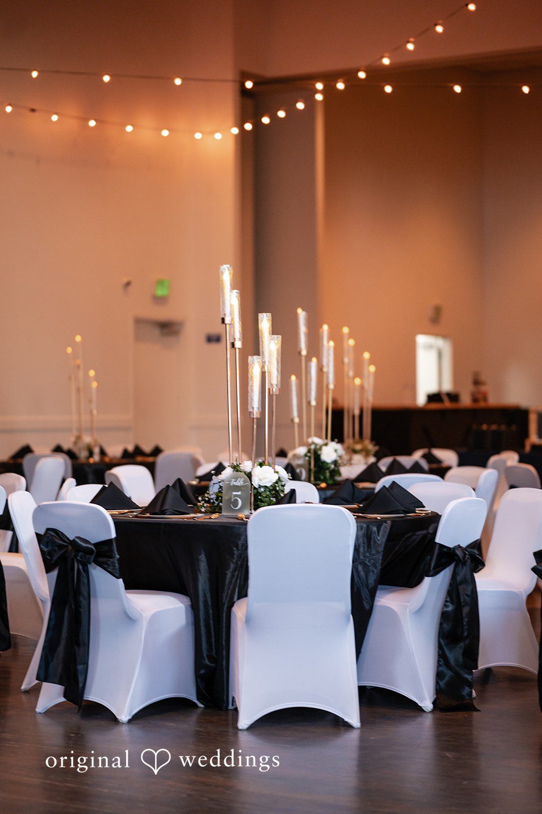 A wedding reception setup showcasing tables with black cloths and chairs with white covers.