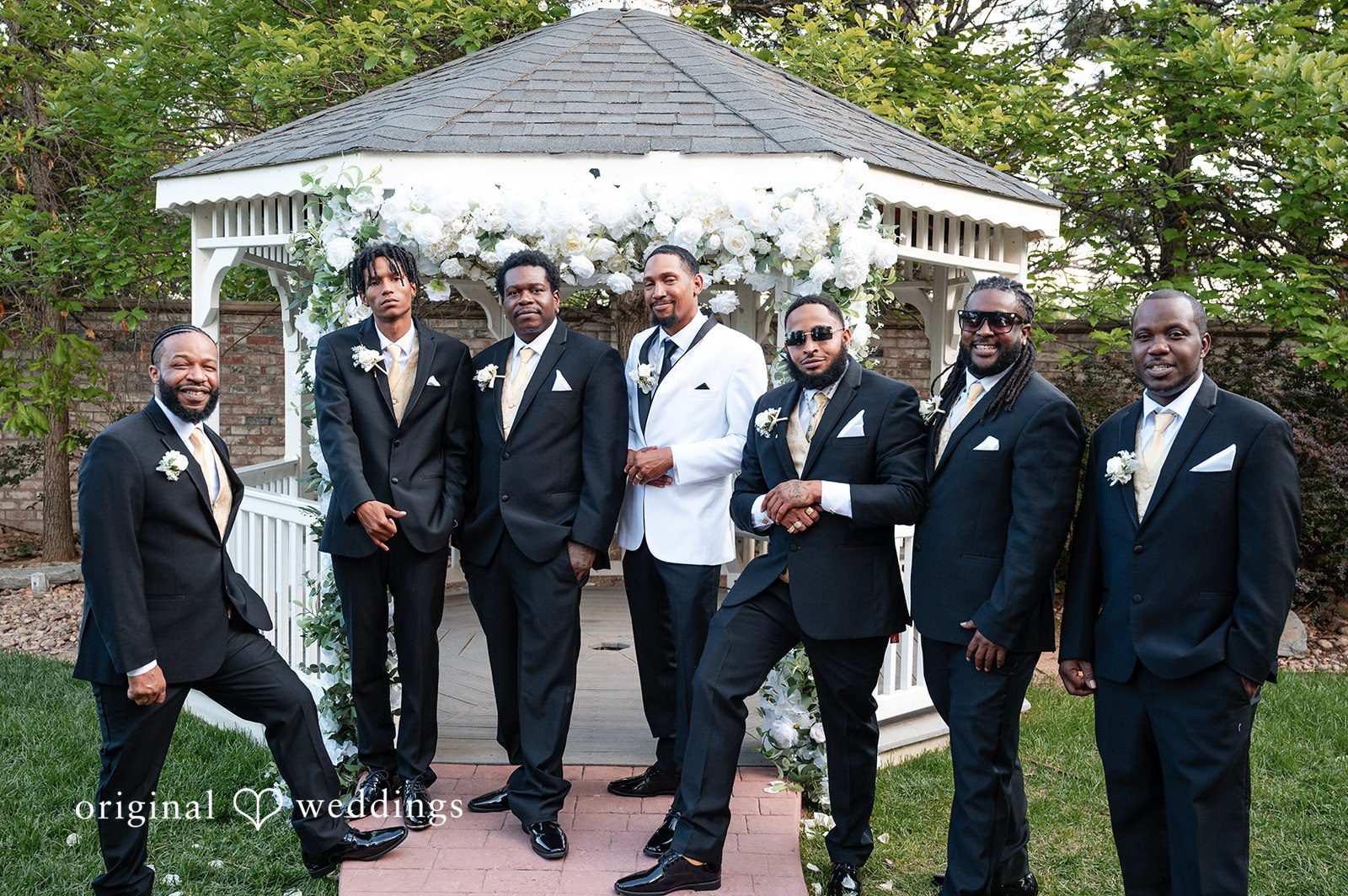 A groom in a white tuxedo posing with five groomsmen in black suits in front of a gazebo.