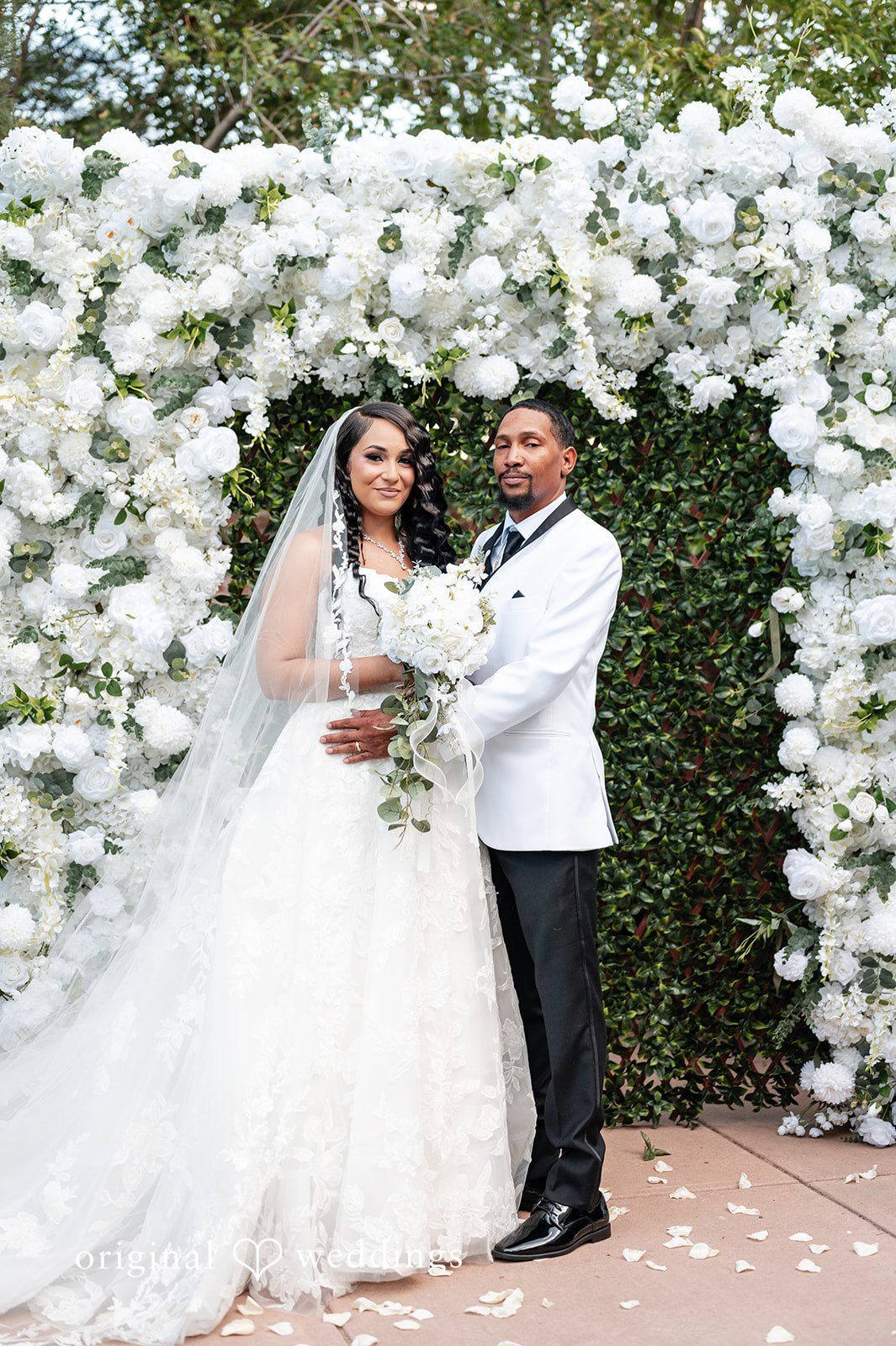 A bride and groom posing together in front of a white floral arch.