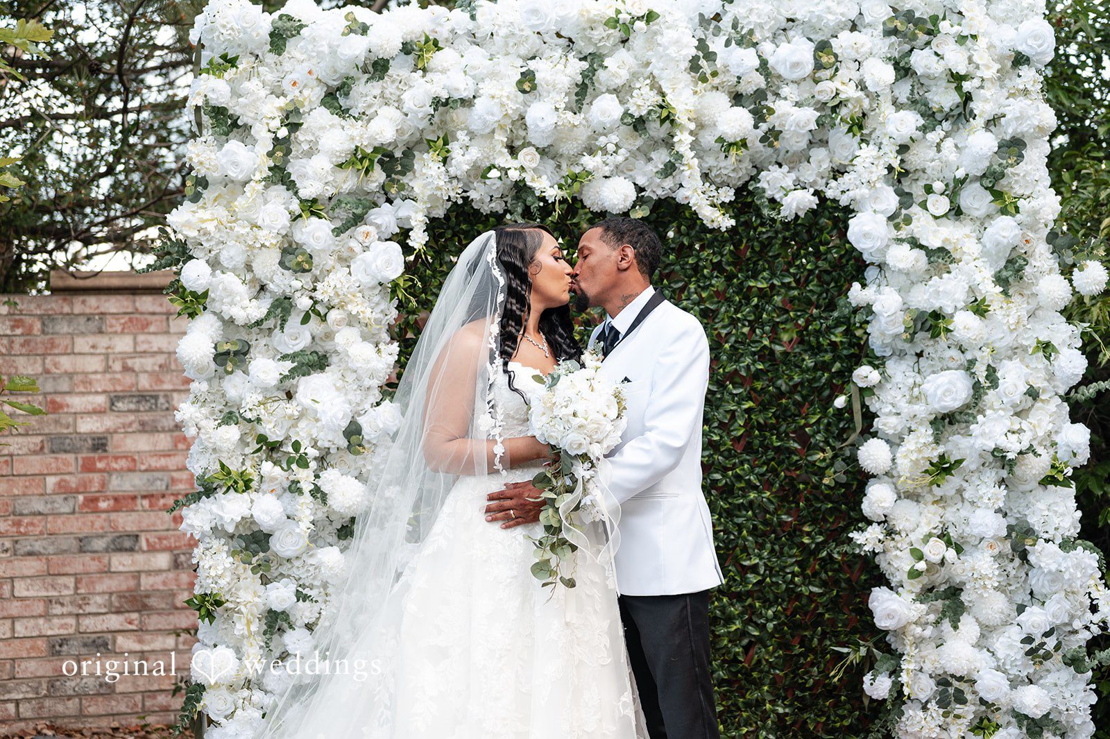 A bride and groom kissing in front of a wall of white flowers.