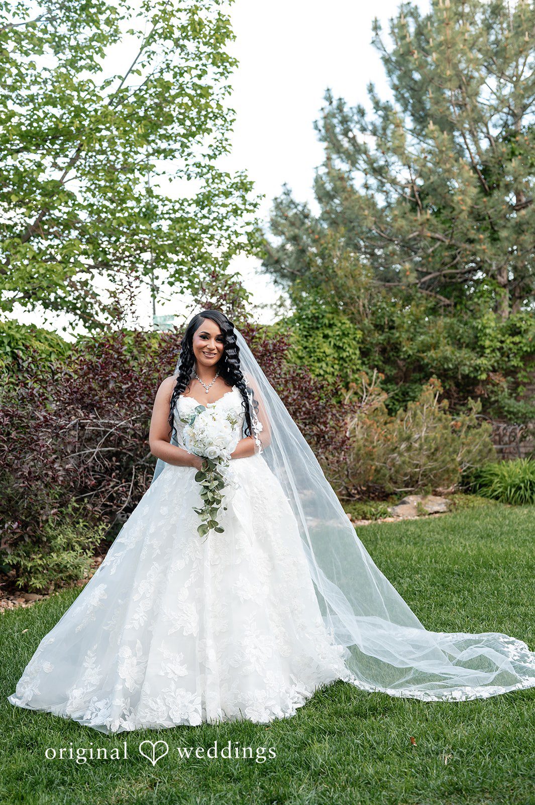 A bride in a white wedding gown and long veil posing on a grassy lawn.