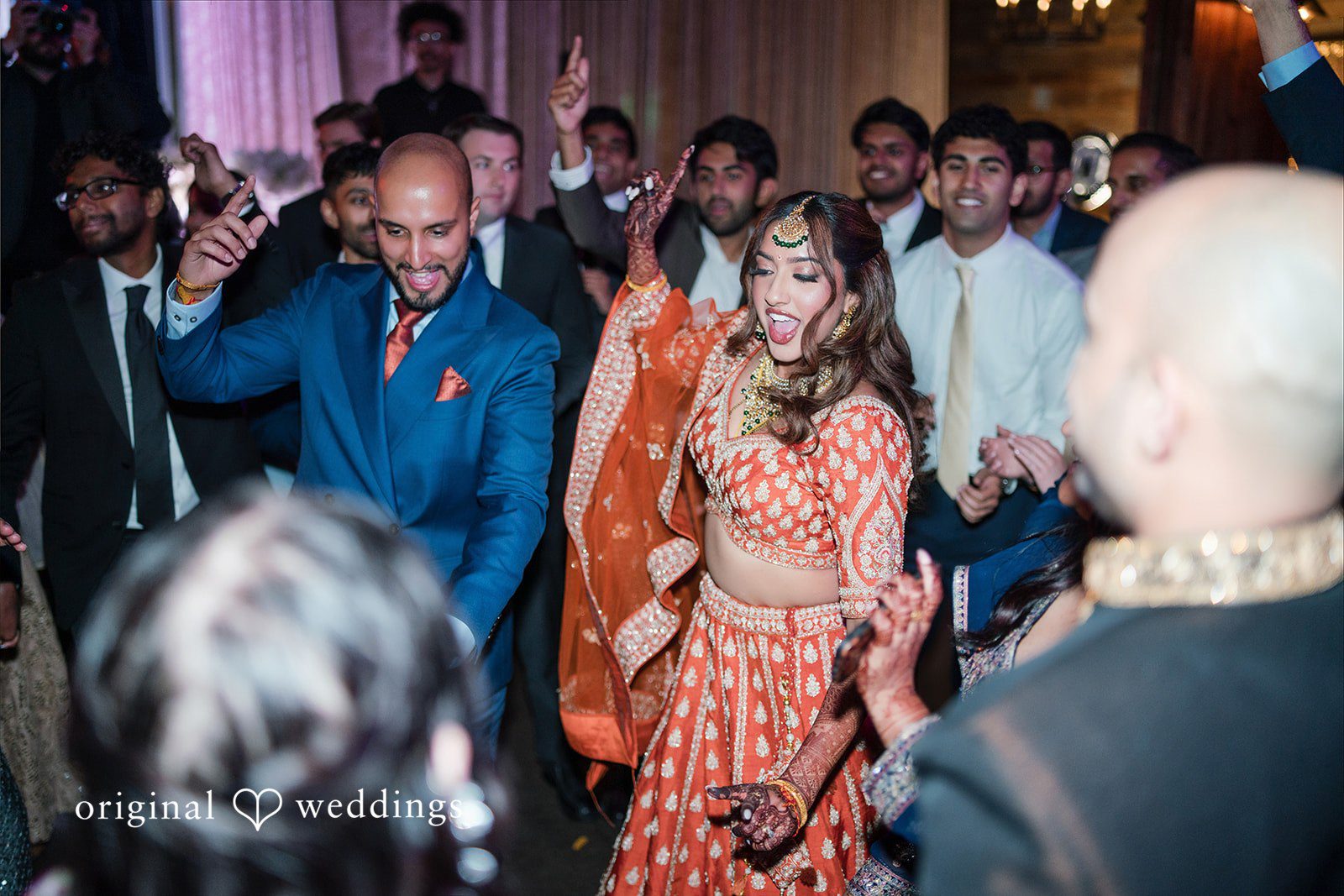 Groom in a blue suit and bride in an orange lehenga dancing energetically amidst a crowd of guests.