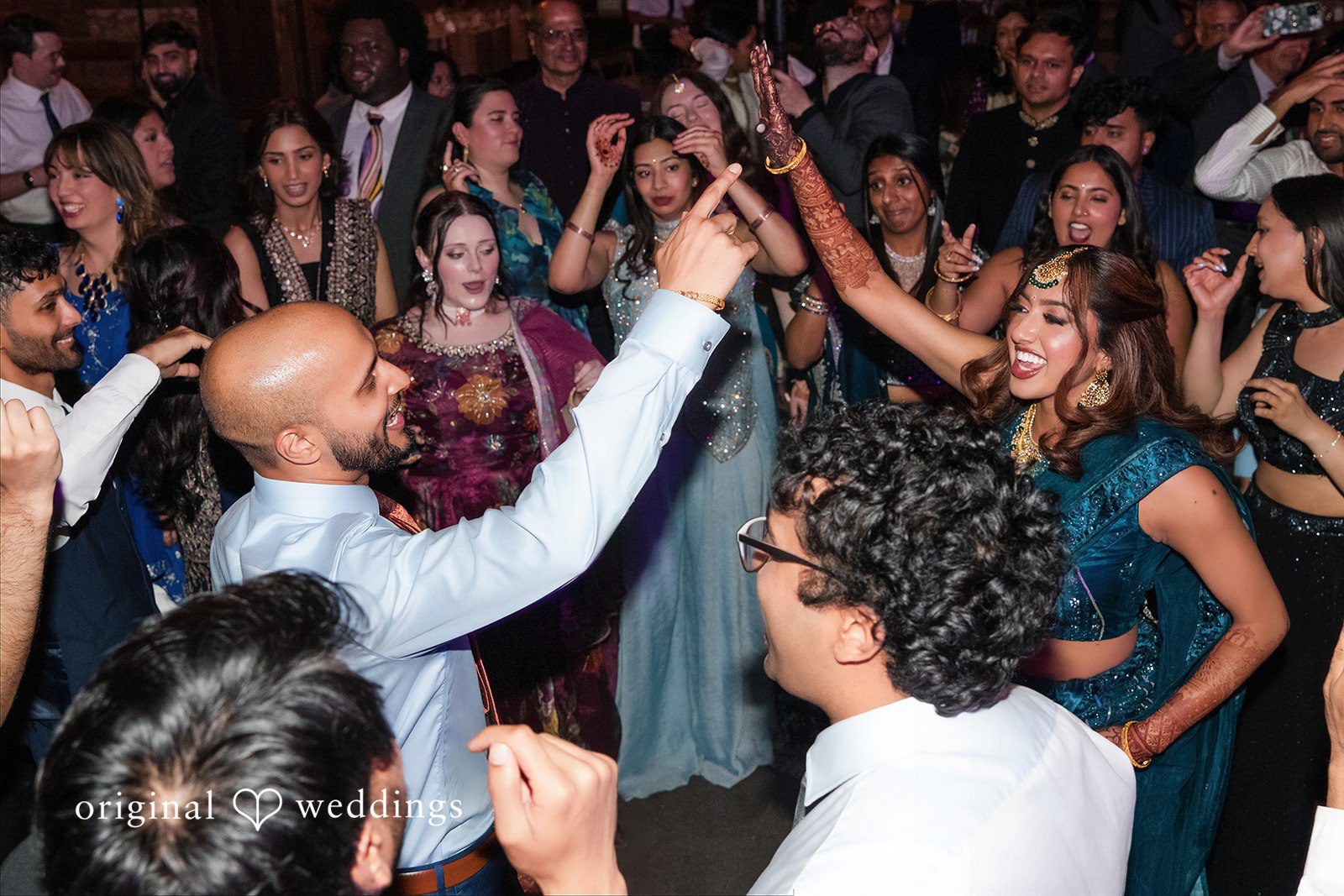 Bride and groom dancing energetically with hands raised amidst a crowded wedding reception dance floor.