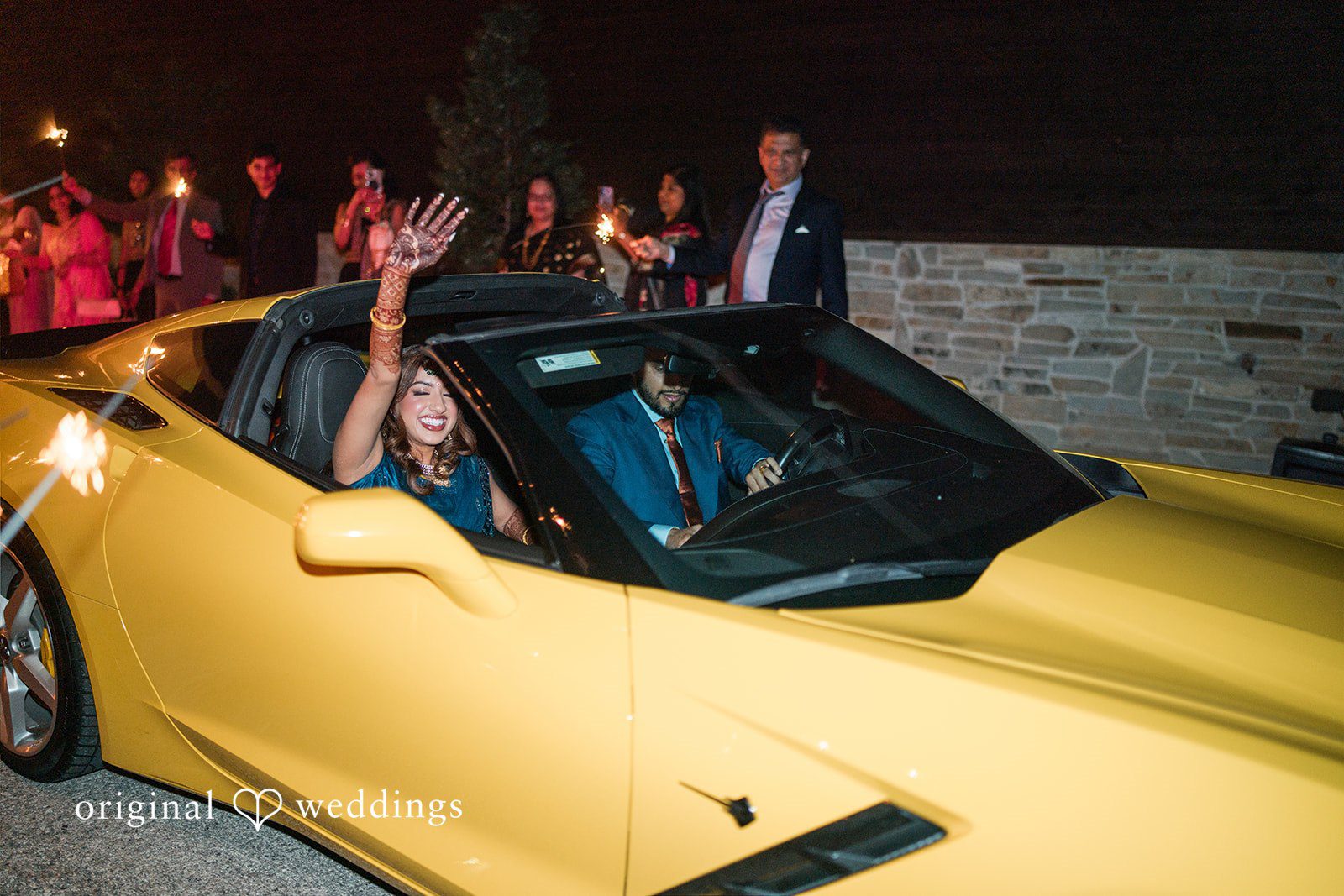 Bride and groom waving from a yellow convertible sports car during their wedding send-off as guests hold sparklers.