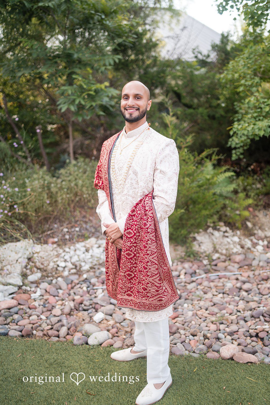 A groom dressed in a traditional cream sherwani and a red shawl posing against a backdrop of greenery.