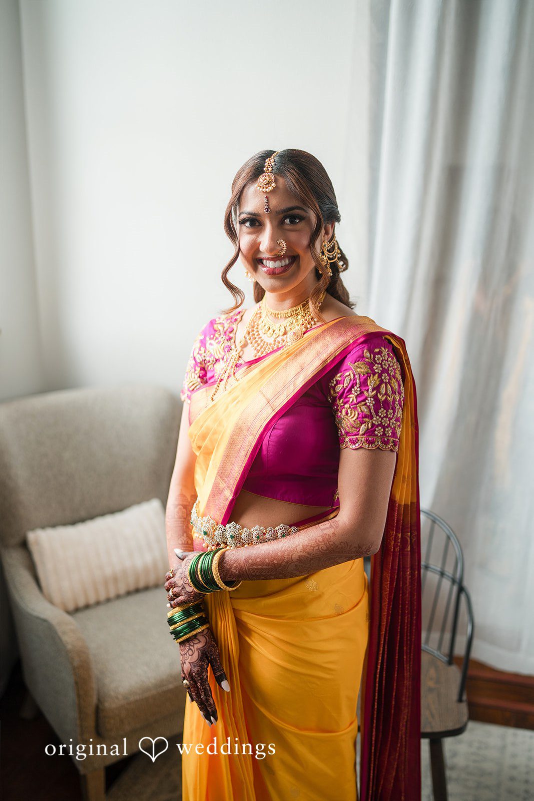 A woman smiling while posing in a yellow and pink saree with a purple blouse.