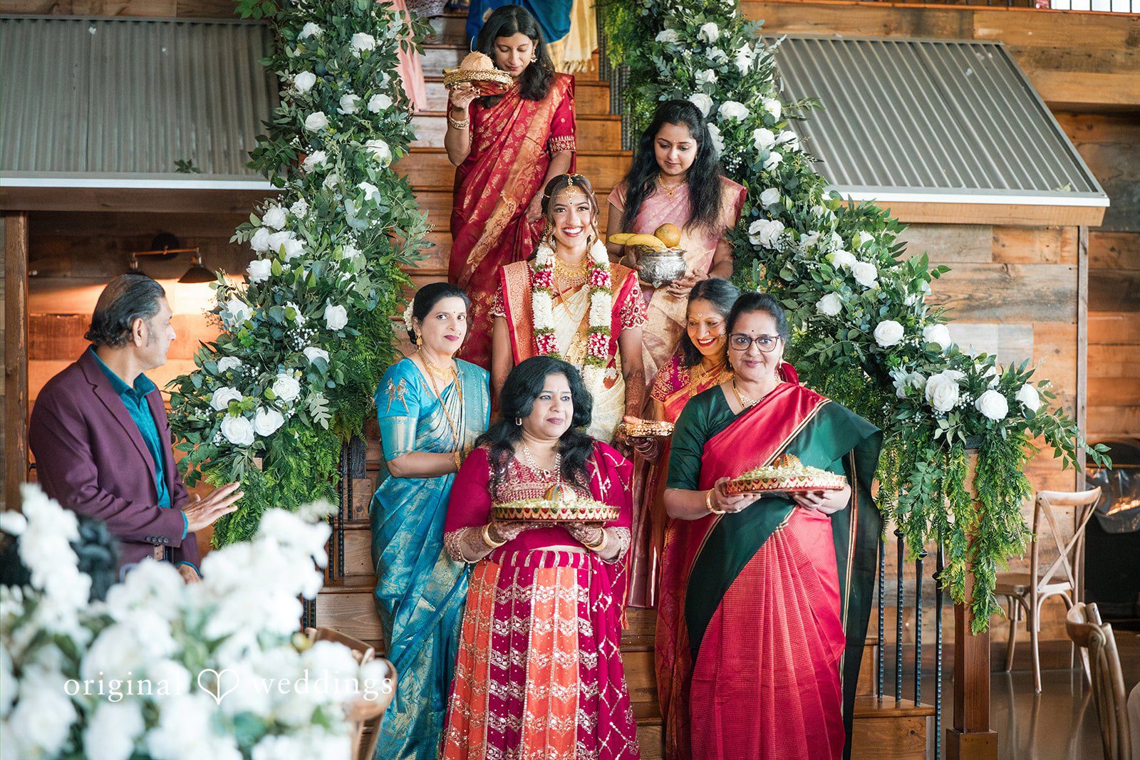 Group photo of wedding guests with bride in colorful traditional attire posed together on decorated stairs.
