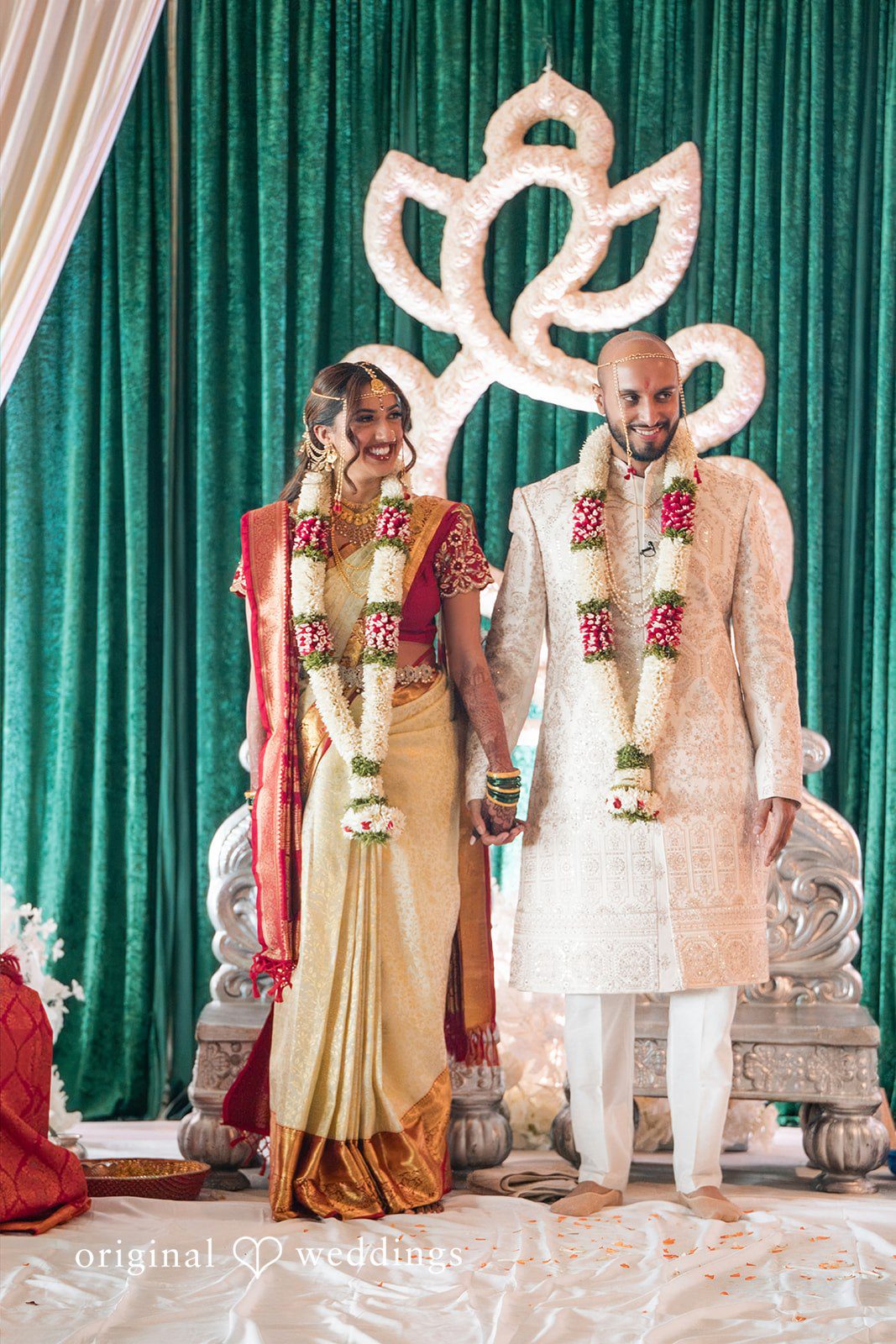 Bride and groom standing together on a decorated stage with a green draped backdrop.