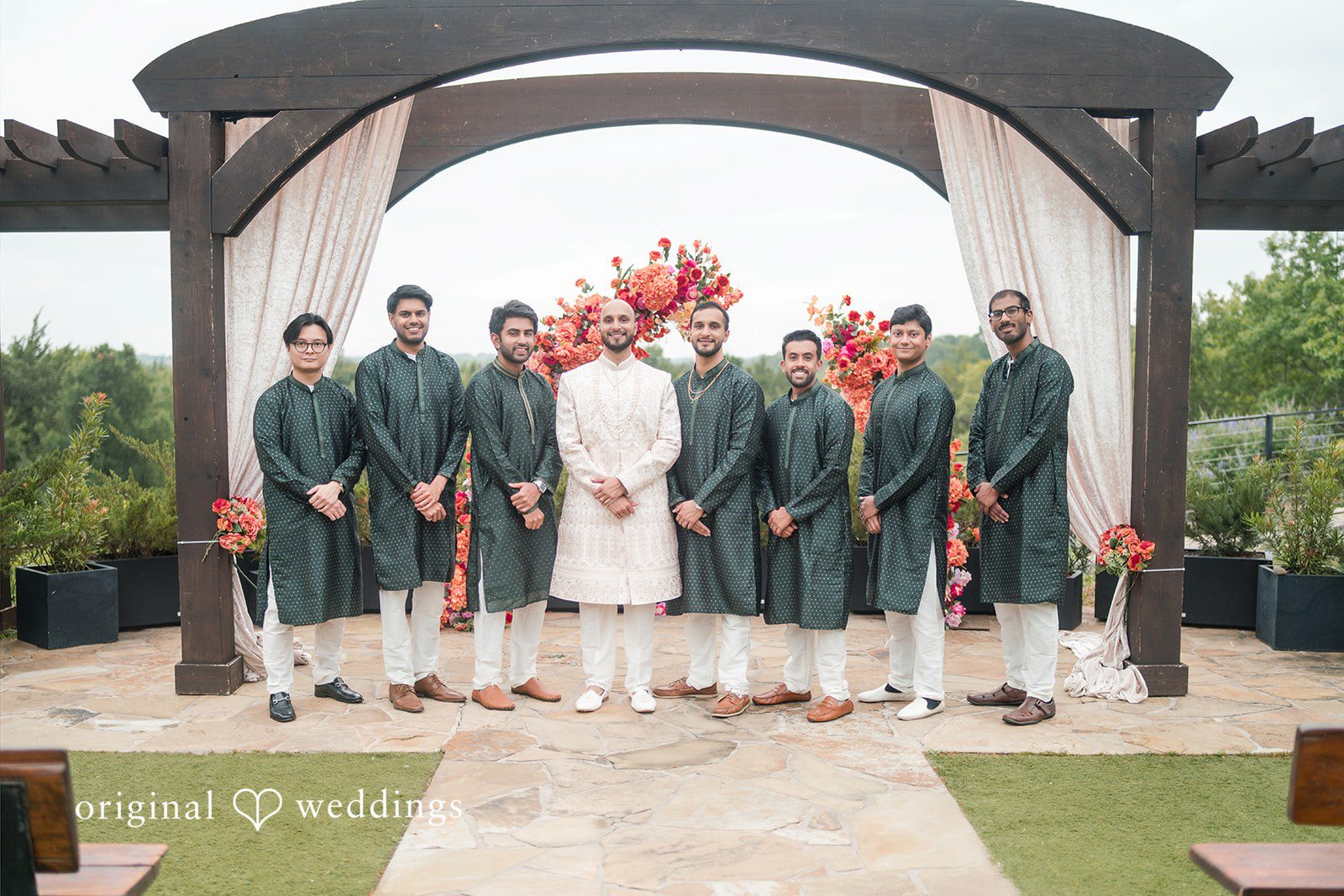 Groom standing with groomsmen wearing matching dark green kurtas under a wooden archway.