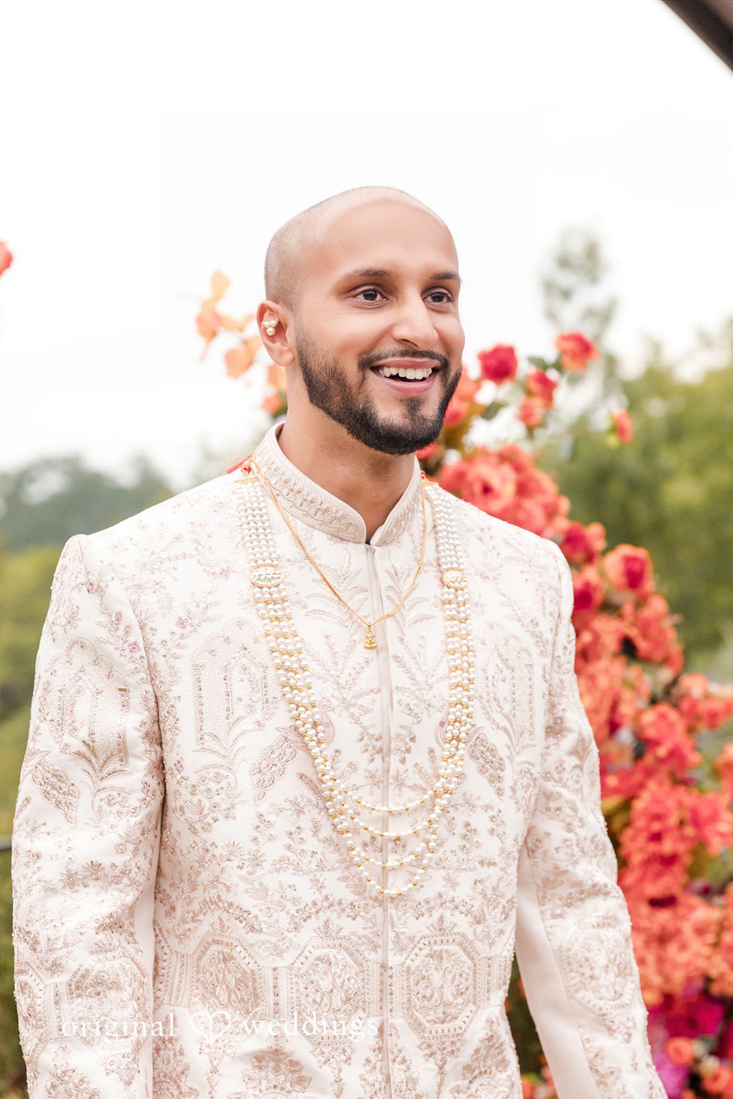 Smiling groom wearing a textured cream sherwani with red floral decorations in the background.