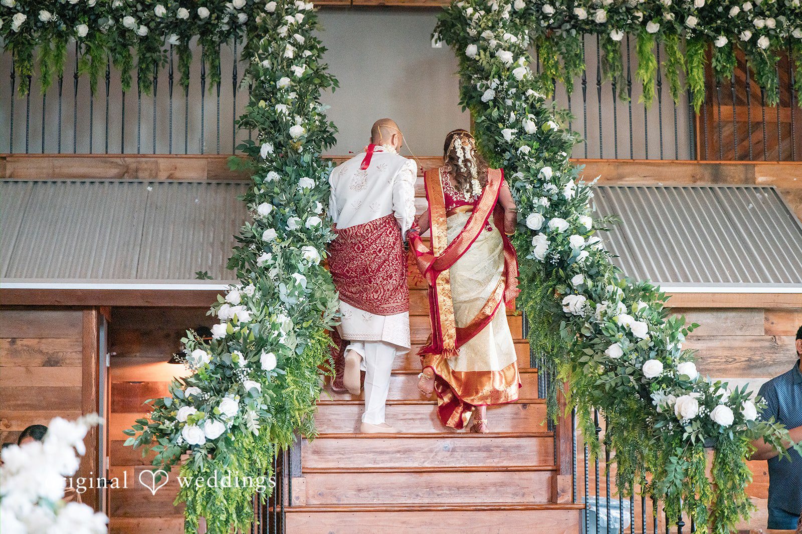 Bride and groom walking down a grand wooden staircase decorated with white floral garlands.