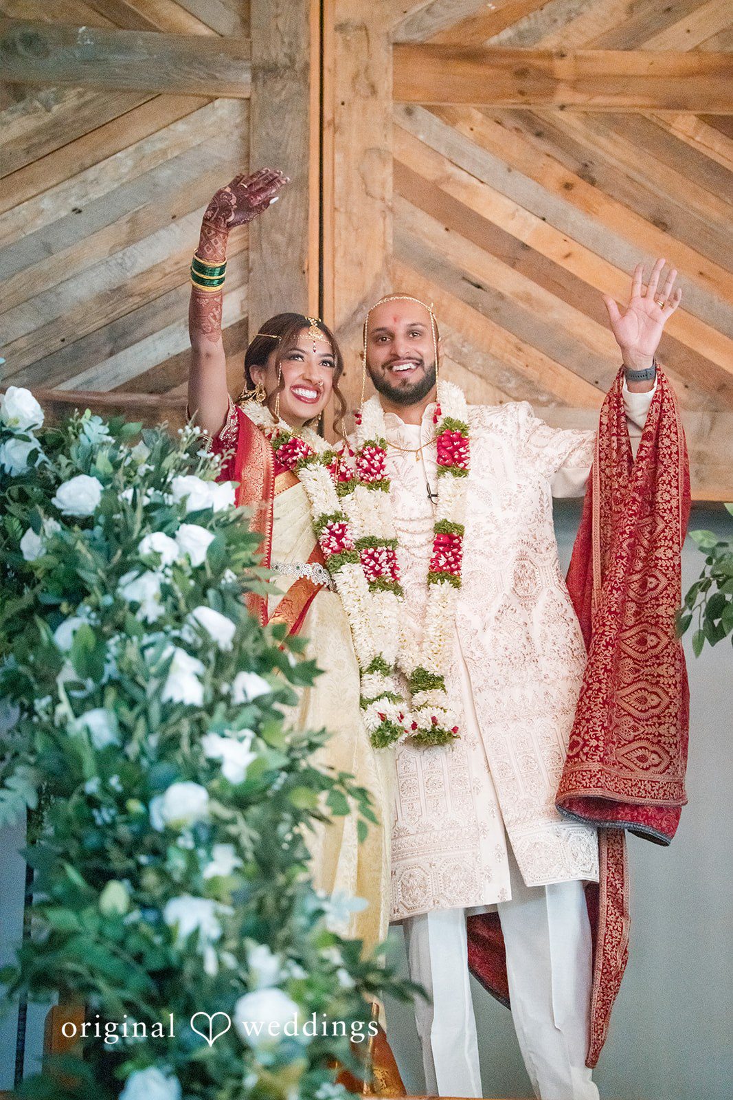 Smiling bride and groom raising their hands in celebration with guest looking on.